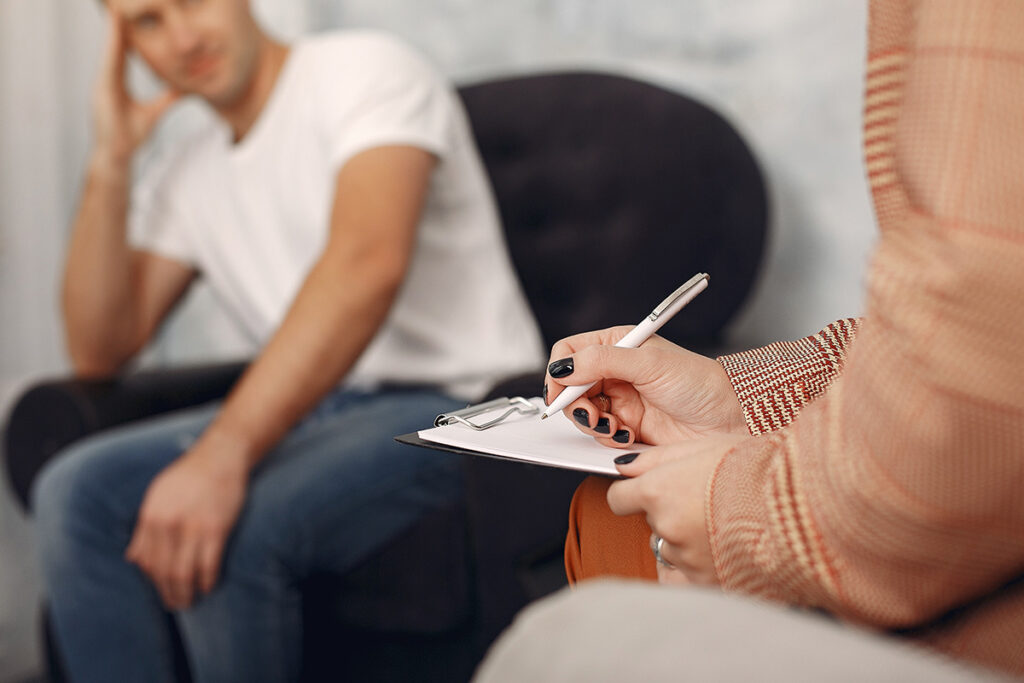 addiction treatment specialist in an office setting writing on clipboard as they explain the connection between anxiety and addiction to a young man.
